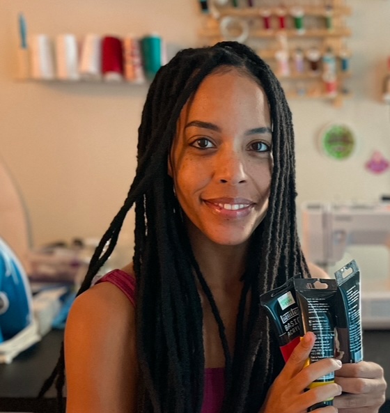 A portrait of Brittany, cosplayer and the sewing teacher of Sewrenity. she is sitting in front of a desk of art supples holding tubes of paint and smiling. African American woman